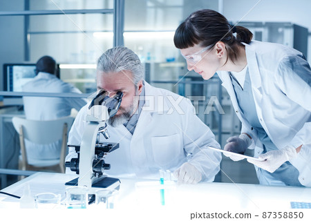 Mature bearded man in white coat examining analysis through the microscope with woman entering data in computer during their teamwork at lab 87358850