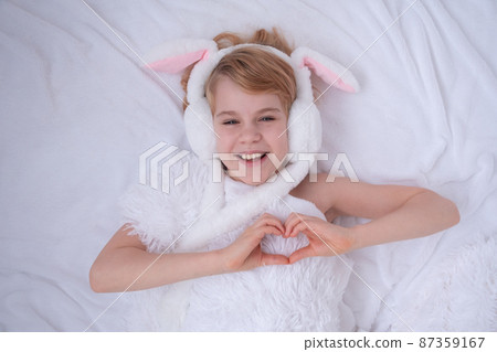 Beautiful little girl plays with rabbit ears. Easter concept. View from above.The child folded the shape of a heart from his fingers. A child on a bed with a fluffy white blanket. Nice and cute Beautiful little girl plays with rabbit ears. Easter concept. View from above.The child folded the shape of a heart from his fingers. A child on a bed with a fluffy white blanket. Nice and cute 87359167