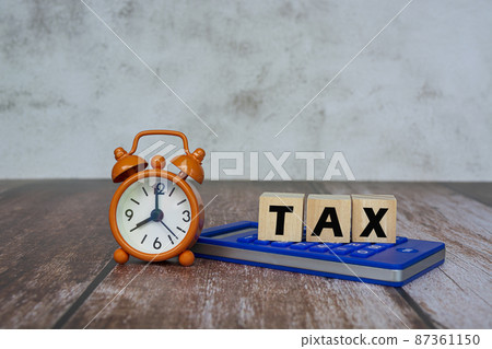 Tax text on wooden cube on a blue calculator with alarm clock set at 8 o'clock. Tax text on wooden cube on a blue calculator with alarm clock set at 8 o'clock. 87361150