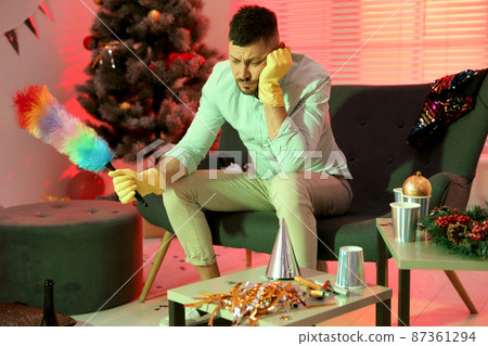 Man with dusting brush suffering from hangover in messy room after New Year party 87361294