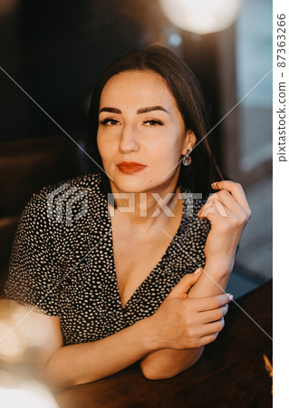 an attractive brunette woman in a black dress at a wooden table in a dark room 87363266