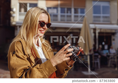 Woman in sunglasses holding camera and looking at it Woman in sunglasses holding camera and looking at it 87363800