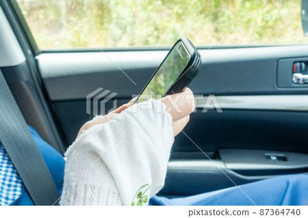 A photo of a woman looking at her smartphone in the passenger seat of a car. Image of smartphone addiction. 87364740