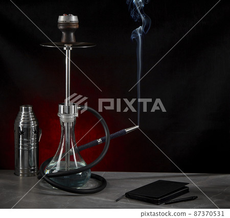 Beautiful modern hookah, from the mouthpiece of which smoke rises, stands on the table, surrounded by a metal protective cap and a diary, on a dark red background. Space for copying. 87370531