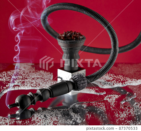 Hookah pipe, curved in a spiral, from the mouthpiece of which smoke flows against the background of a ceramic hookah bowl with a small stand on a scarlet background with reflections. Hookah pipe, curved in a spiral, from the mouthpiece of which smoke flows against the background of a ceramic hookah bowl with a small stand on a scarlet background with reflections. 87370533