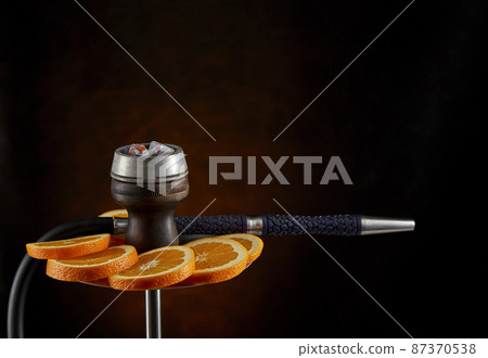 Modern hookah decorated with orange slices on a blurry dark red background. The culture of serving shisha. 87370538