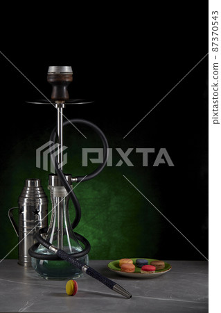 Beautiful modern hookah standing on a table surrounded by a metal protective cap and a plate with colorful macaroons on a dark green background. Space for copying. Vertical location. 87370543