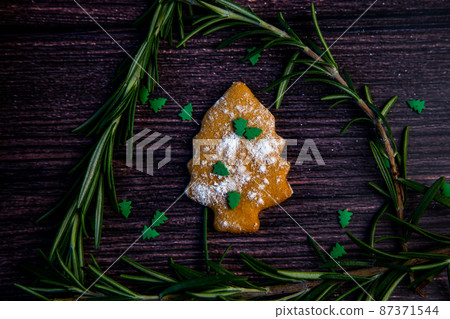Cookies in the form of a Christmas tree, sprinkled with powdered sugar and small Christmas trees on top, lies on a dark wooden background surrounded by grained rosemary. High quality photo Cookies in the form of a Christmas tree, sprinkled with powdered sugar and small Christmas trees on top, lies on a dark wooden background surrounded by grained rosemary. High quality photo 87371544