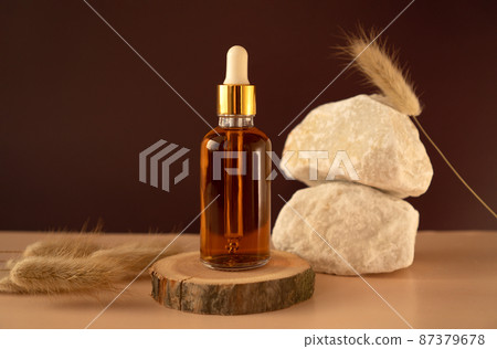 Transparent glass cosmetic dropper standing on wooden podium near stones and dry flowers on dark beige background. Beauty composition. Concept of natural and original skincare products presentation. Transparent glass cosmetic dropper standing on wooden podium near stones and dry flowers on dark beige background. Beauty composition. Concept of natural and original skincare products presentation. 87379678