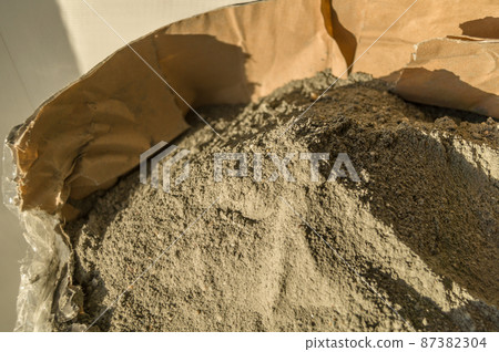 Grey sand-concrete dry mix in an open paper bag. Concrete dry powder for leveling walls and floors. Repair in the house with your own hands. DIY. Selective focus Grey sand-concrete dry mix in an open paper bag. Concrete dry powder for leveling walls and floors. Repair in the house with your own hands. DIY. Selective focus 87382304