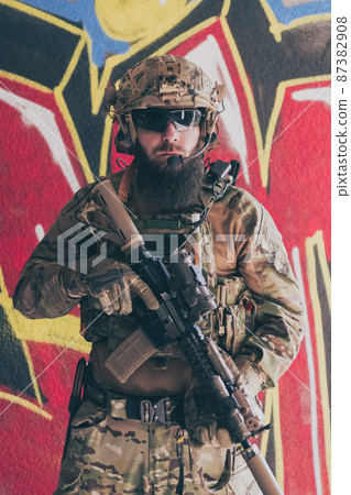 A bearded soldier in a special forces uniform stands in front of a drawn wall with a rifle in his hand. Selective focus  87382908