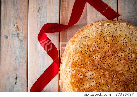 Traditional Russian thin pancakes closeup on a light wooden background and a red ribbon. Traditional Russian thin pancakes closeup on a light wooden background and a red ribbon. 87386487