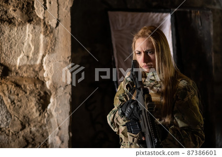 Caucasian woman in army uniform holding a machine gun and standing near an open coffin.  87386601