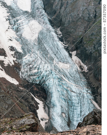 Vertical mountain landscape with blue long vertical glacier tongue with cracks among rocks. Aerial view to large glacier with icefall. Nature texture of mountain glacier with fissures close up. Vertical mountain landscape with blue long vertical glacier tongue with cracks among rocks. Aerial view to large glacier with icefall. Nature texture of mountain glacier with fissures close up. 87387090
