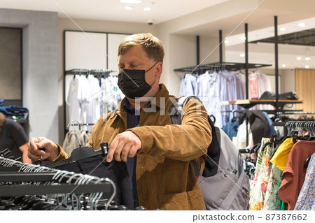 A man in a shopping mall in a protective black mask chooses clothes of his size A man in a shopping mall in a protective black mask chooses clothes of his size 87387662