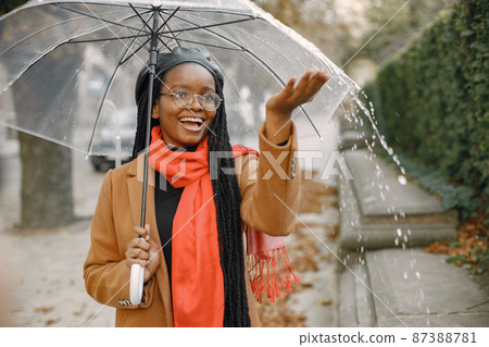 Young black woman in a coat standing under a transparent umbrella outside 87388781