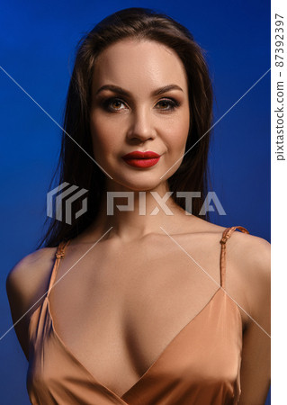 Brunette girl with bright make-up and deep neckline, in silk beige dress. She is smiling, posing against blue studio background. Close up 87392397