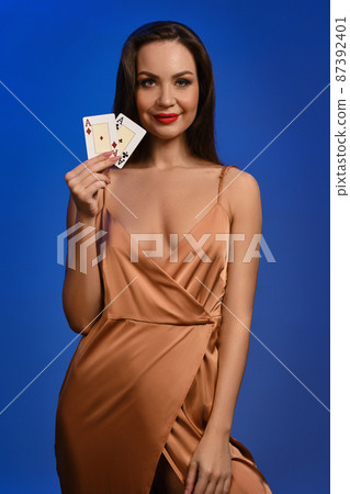 Brunette lady in silk golden dress. She is smiling, showing two playing cards, posing on blue background. Poker, casino. Close-up 87392401