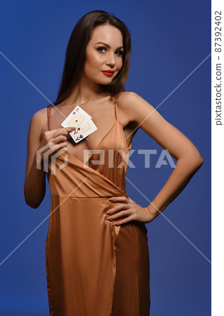 Brunette girl in silk golden dress. She smiling, showing two aces, posing against blue background. Gambling, poker, casino. Close-up Brunette girl in silk golden dress. She smiling, showing two aces, posing against blue background. Gambling, poker, casino. Close-up 87392402
