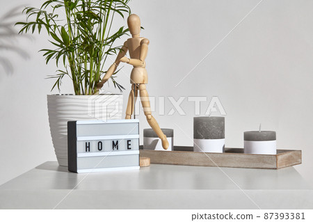 Table, gray different sized candles on wooden stand, figurine of human, lamp with inscription home, green plant in pot. Isolated on white. Close up 87393381