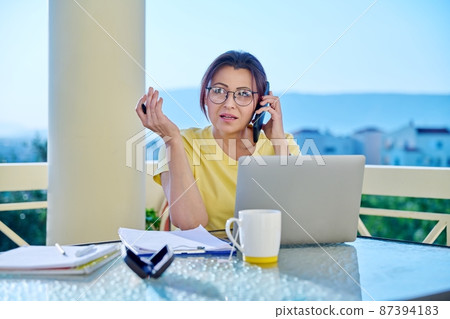 Middle-aged woman working in home office on terrace, talking on phone Middle-aged woman working in home office on terrace, talking on phone 87394183