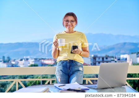 Middle-aged woman working at home on terrace, relaxing with mug of tea and smartphone 87394184