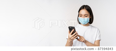 Health and covid-19 concept. Young asian woman in medical mask using mobile phone, typing on smartphone, standing against white background Health and covid-19 concept. Young asian woman in medical mask using mobile phone, typing on smartphone, standing against white background 87394843