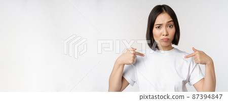 Young asian woman pointing at herself with disbelief, being chosen, surprised by her candidature, standing over white background Young asian woman pointing at herself with disbelief, being chosen, surprised by her candidature, standing over white background 87394847