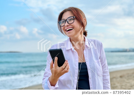 Happy middle aged woman on sea vacation enjoying in headphones with smartphone 87395402