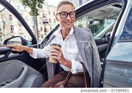 Starting day with coffee. Beautiful middle aged business woman arrived at work, she is getting out of her modern car and smiling Starting day with coffee. Beautiful middle aged business woman arrived at work, she is getting out of her modern car and smiling 87399463