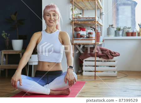 Smiling woman with smartwatch sits in lotus pose at home Smiling woman with smartwatch sits in lotus pose at home 87399529