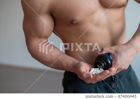 Unrecognizable bodybuilder man drinks vitamin capsules. A large male muscular torso pours a handful of sports supplements from the jar. Unrecognizable bodybuilder man drinks vitamin capsules. A large male muscular torso pours a handful of sports supplements from the jar. 87400445