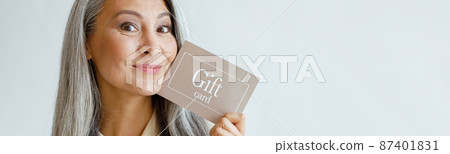 Joyful grey haired Asian woman holds gift card posing on light grey background Joyful grey haired Asian woman holds gift card posing on light grey background 87401831
