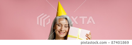 Happy mature Asian lady with yellow party hat holds gift box on pink background 87401869