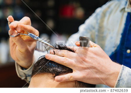 hairdresser makes a haircut for a brunet man with a scissors in a barbershop 87402639