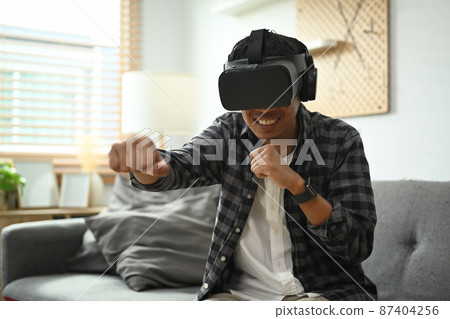 Man wearing virtual reality headset playing simulation boxing game in living room. 87404256