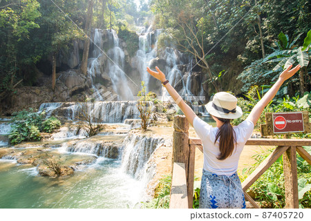 Back view of young woman tourist raised her arms and looking to the beautiful landscape of Kuang Si falls an iconic waterfalls in the jungle park nearly Luang Prabang town, in north central Laos. Back view of young woman tourist raised her arms and looking to the beautiful landscape of Kuang Si falls an iconic waterfalls in the jungle park nearly Luang Prabang town, in north central Laos. 87405720
