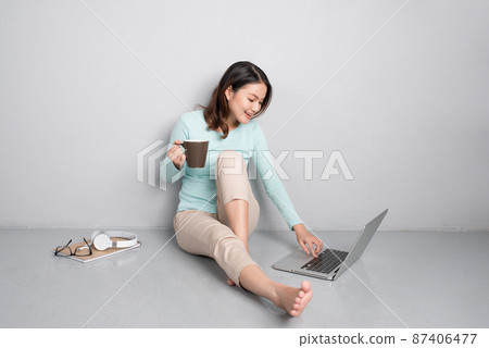Happy casual beautiful asian woman working on a laptop sitting on floor at home. 87406477