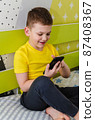 The boy plays on a smartphone while sitting in his room on the bed. 87408367