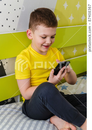 The boy plays on a smartphone while sitting in his room on the bed. 87408367