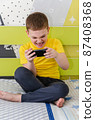 The boy plays on a smartphone while sitting in his room on the bed. 87408368