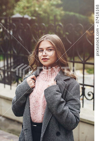 Fashionable and beautiful young woman 16-20 years old, dressed in an pink knitted sweater, glasses, dark skirt and a gray classic trench coat. 87416168