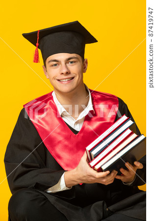 Portrait of young smiling man, student in graduation cap and gown with books posing isolated over yellow studio background 87419747