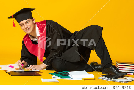 Portrait of young smiling man, student in graduation cap and gown lying on floor and doing homework isolated over yellow studio background Portrait of young smiling man, student in graduation cap and gown lying on floor and doing homework isolated over yellow studio background 87419748
