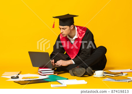Portrait of young man, student in graduation cap and gown typing project on laptop isolated over yellow studio background Portrait of young man, student in graduation cap and gown typing project on laptop isolated over yellow studio background 87419813
