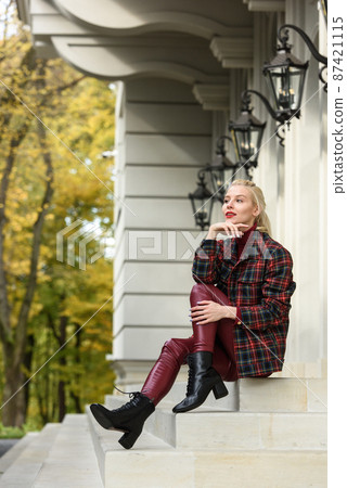 A beautiful, modern, fashionable blonde girl with a red lipstick posing outdoors . red leather leggings, turtleneck and checkered jacket. fit figure. A beautiful, modern, fashionable blonde girl with a red lipstick posing outdoors . red leather leggings, turtleneck and checkered jacket. fit figure. 87421115