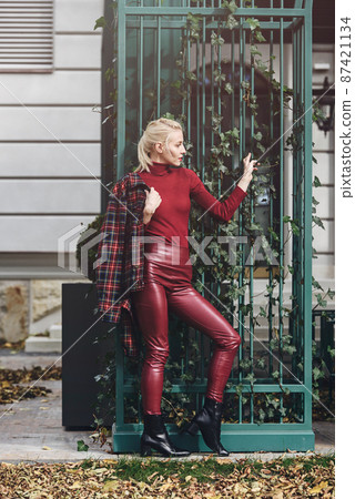 fashionable blonde girl with a red lipstick posing outdoors . Dressed in a red leather leggings, turtleneck and checkered jacket. fit figure fashionable blonde girl with a red lipstick posing outdoors . Dressed in a red leather leggings, turtleneck and checkered jacket. fit figure 87421134