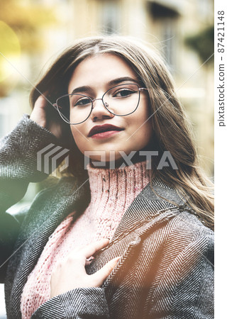 Fashionable portrait of a beautiful young woman 16-20 years old, dressed in an pink knitted sweater, glasses and a gray classic trench coat. 87421148