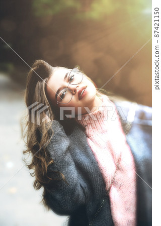 Fashionable portrait of a beautiful young woman 16-20 years old, dressed in an pink knitted sweater, glasses and a gray classic trench coat. 87421150