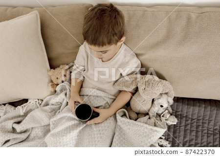 Sick little boy drinking hot tea on the bed at home. Unwell, illness child wrapped in a blanket, with mug in his room. Flu season. Interior and clothes in natural earth colors. Cozy environment. Sick little boy drinking hot tea on the bed at home. Unwell, illness child wrapped in a blanket, with mug in his room. Flu season. Interior and clothes in natural earth colors. Cozy environment. 87422370